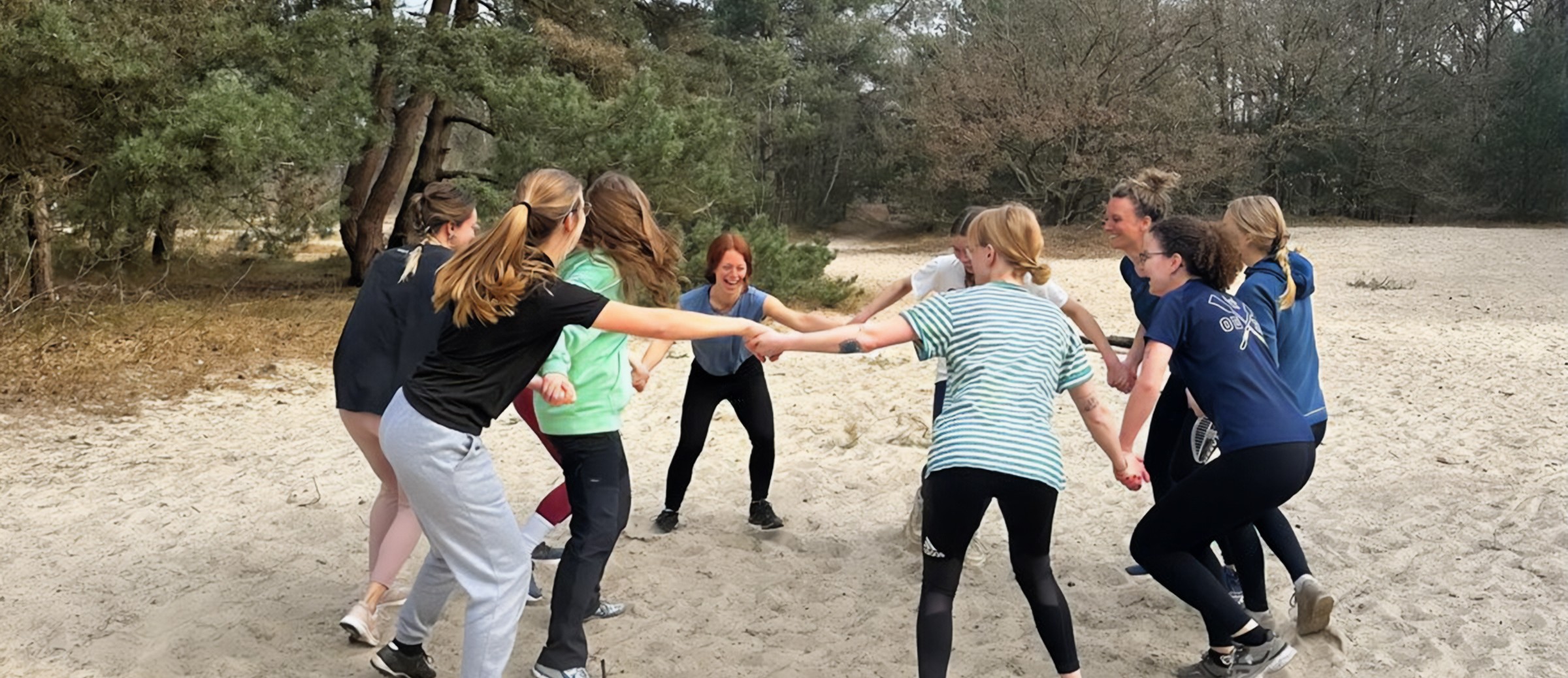 Groep vrouwen in sportkleding vormt een kring op een zandpad omgeven door bomen.