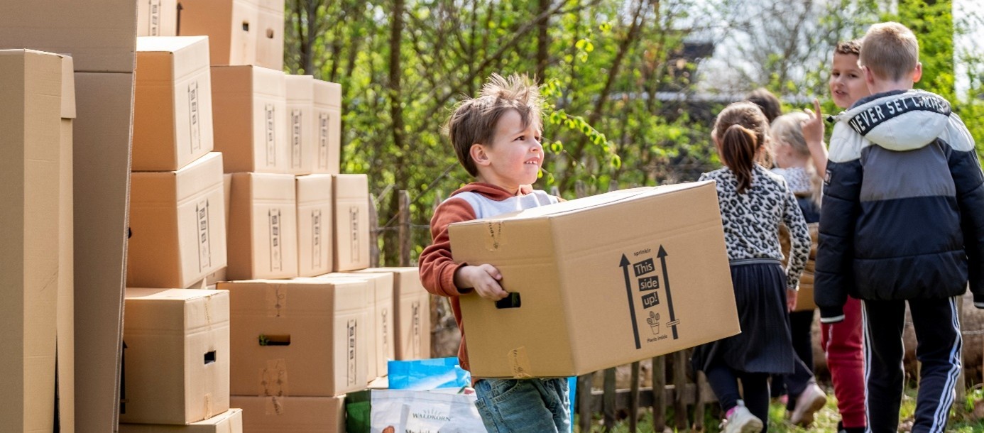 Kind tilt doos buiten naast stapel dozen, met kinderen op de achtergrond.