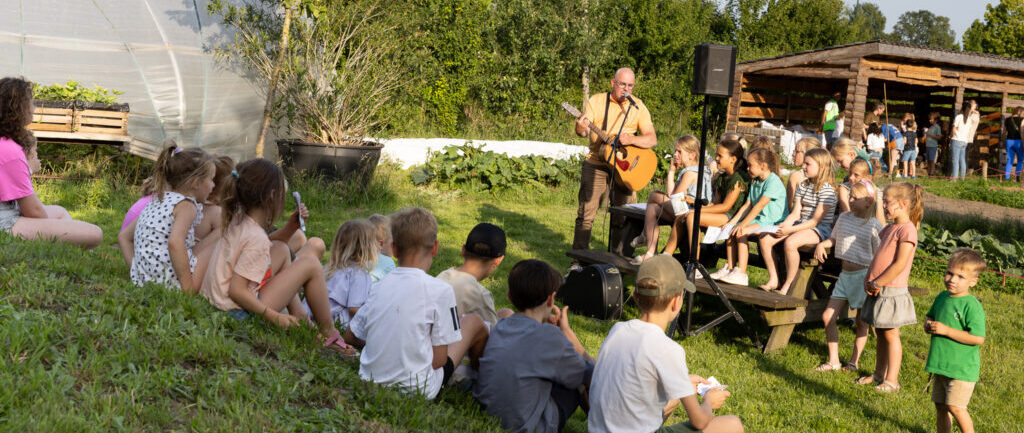 Muzikant met gitaar speelt voor kinderen die op grasveld zitten bij een kas en houten schuur.