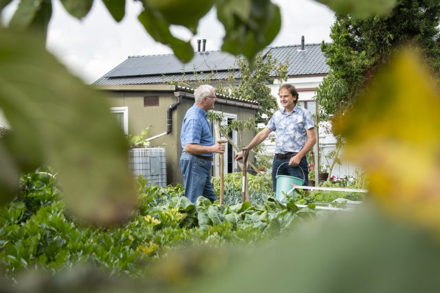 Twee mannen praten in een tuin vol groen, met een huis op de achtergrond.