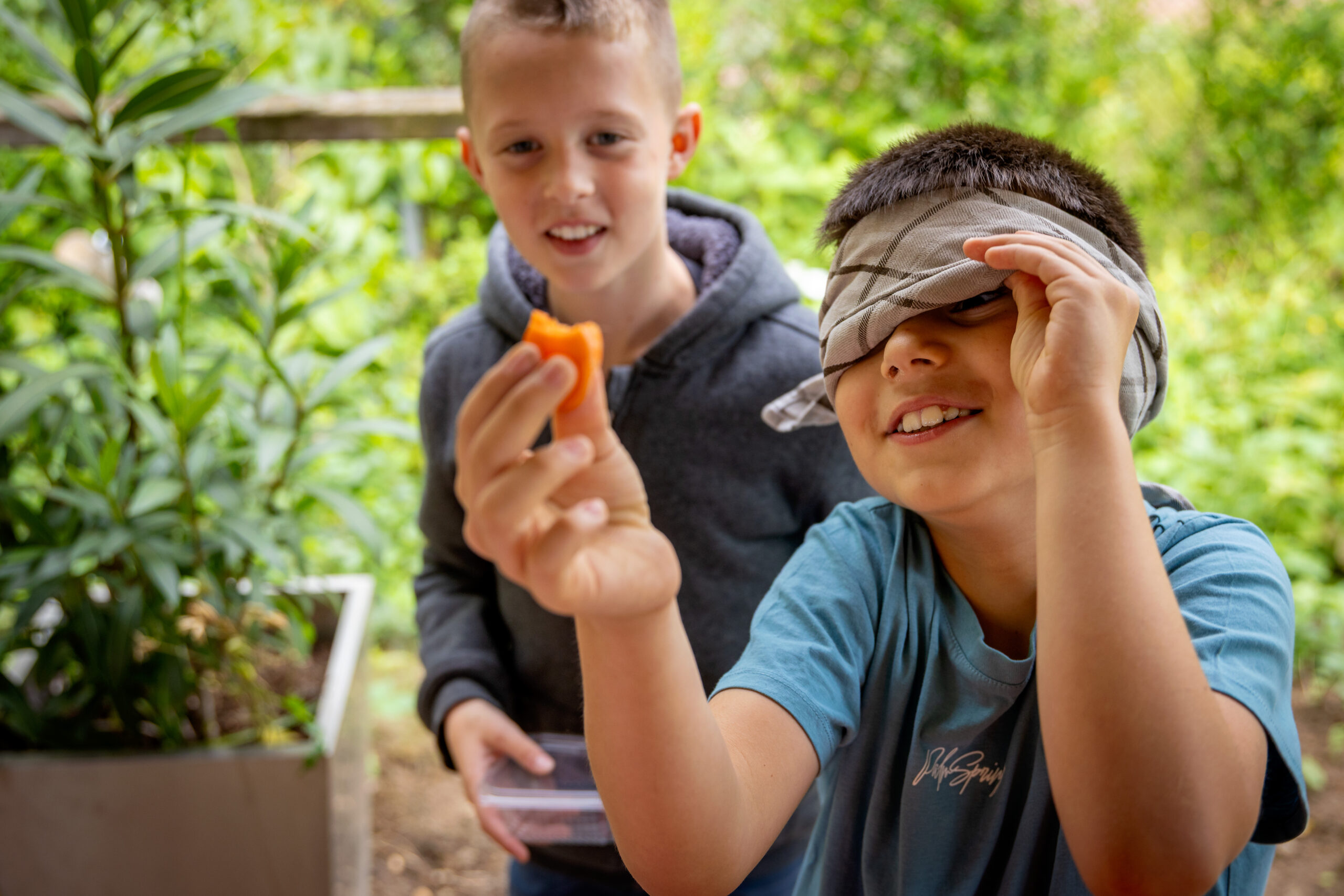 Twee kinderen, één geblinddoekt, houden snack vast in groene buitenomgeving.