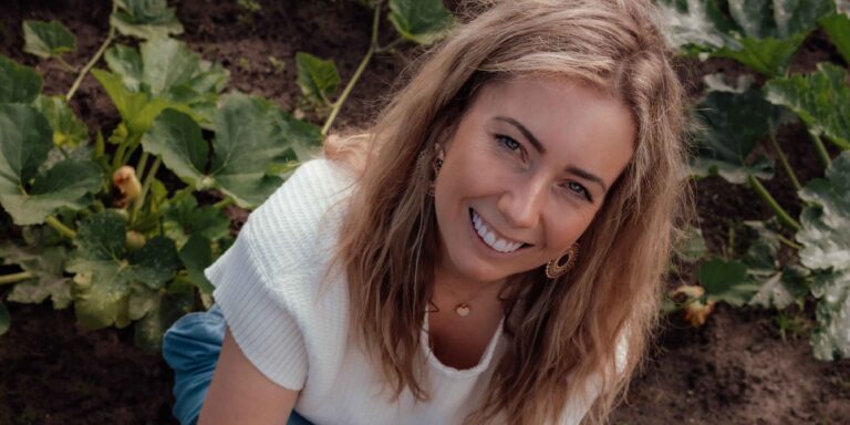 Vrouw glimlachend in een tuin met groene planten op de achtergrond.