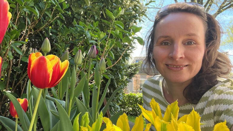Vrouw glimlacht naast kleurrijke tulpen en groene bladeren in een zonnige tuin.
