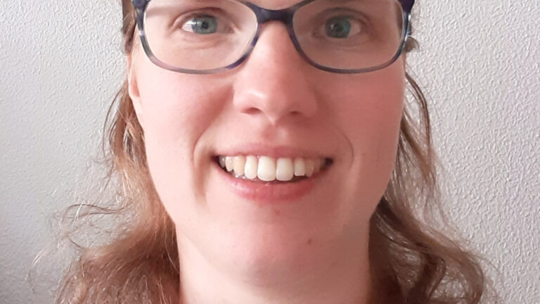 Close-up van glimlachende persoon met bril tegen een lichtgekleurde muur.