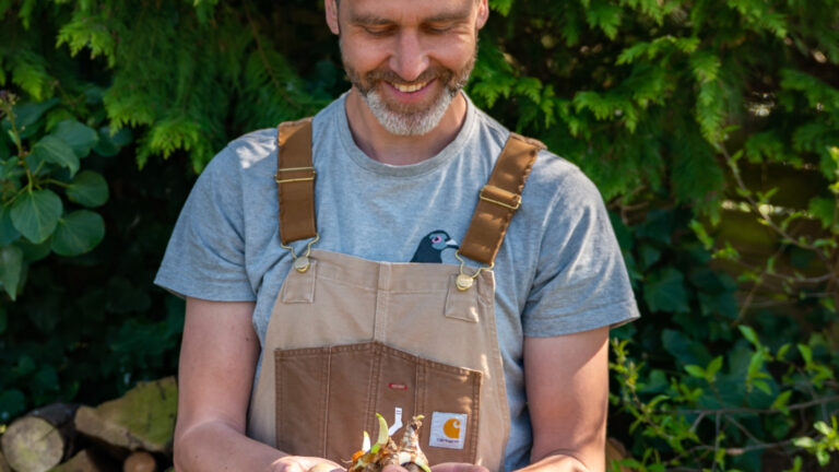 Man in tuinwerk outfit houdt kleine bloembollen vast, omgeven door groen.