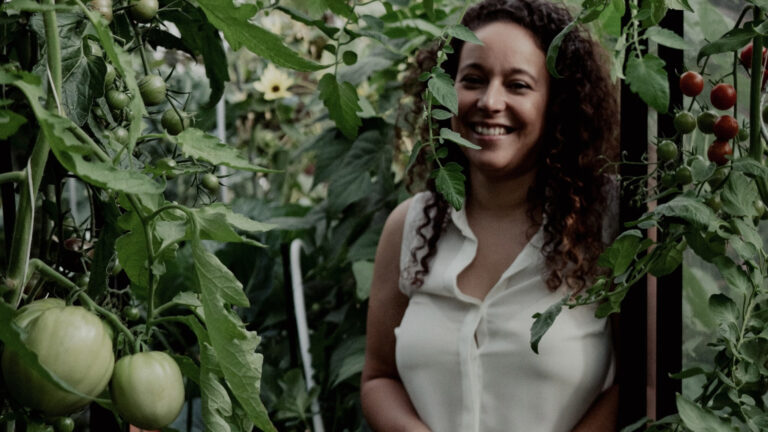 Vrouw in kas omringd door groene en rode tomatenplanten, glimlachend naar de camera.