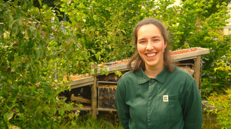 Vrouw in groene outfit lacht in een groene tuin met een insectenhotel op de achtergrond.