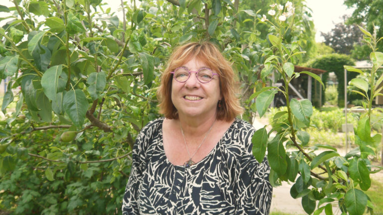 Vrouw met bril lacht voor boom met groene bladeren in tuinomgeving.