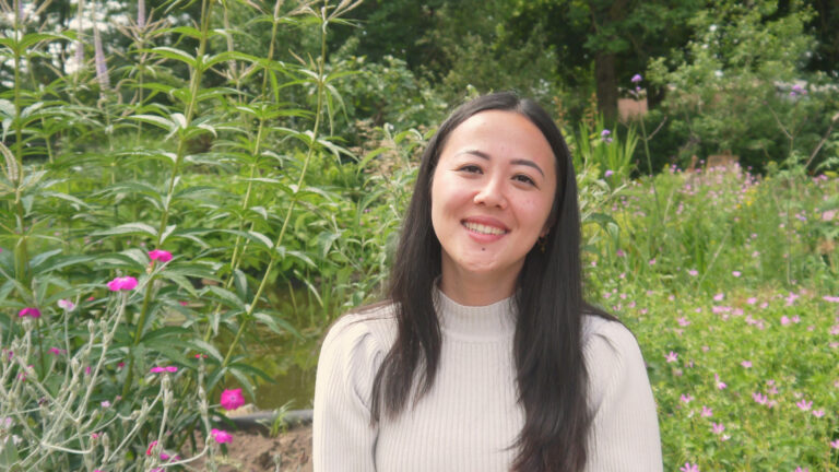 Vrouw glimlacht in een tuin met groene planten en roze bloemen op de achtergrond.