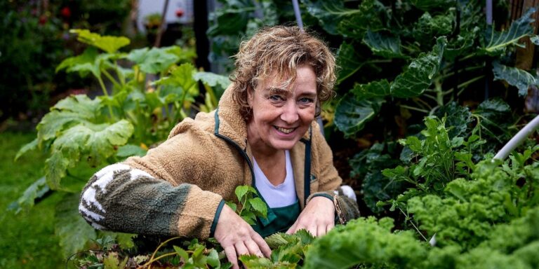 Vrouw met krullend haar werkt glimlachend in een weelderige groentetuin.