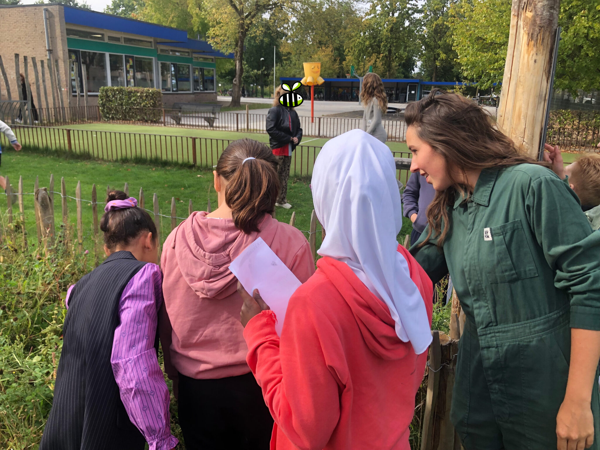 Kinderen en volwassenen onderzoeken planten bij een schoolplein met gebouw en bomen op de achtergrond.