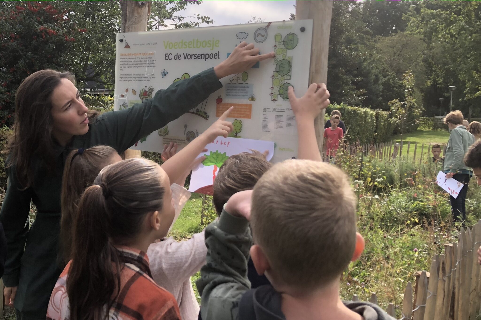 Docent wijst iets aan op een infobord, omringd door nieuwsgierige kinderen in een voedselbos.
