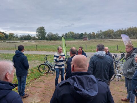 Een groep mensen luistert naar een spreker buiten op een herfstdag, fietsen op de achtergrond.