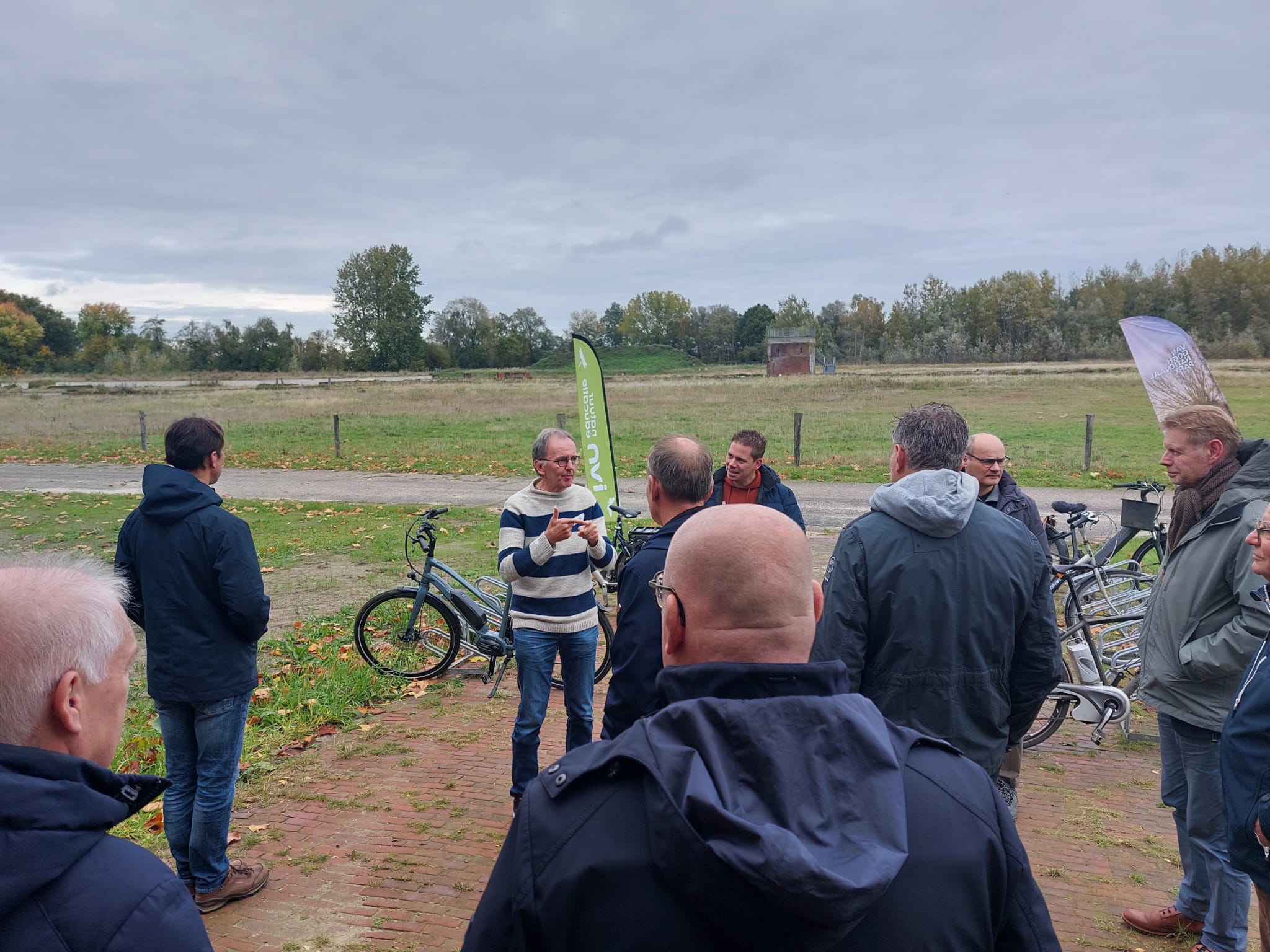 Een groep mensen luistert naar een spreker buiten op een herfstdag, fietsen op de achtergrond.
