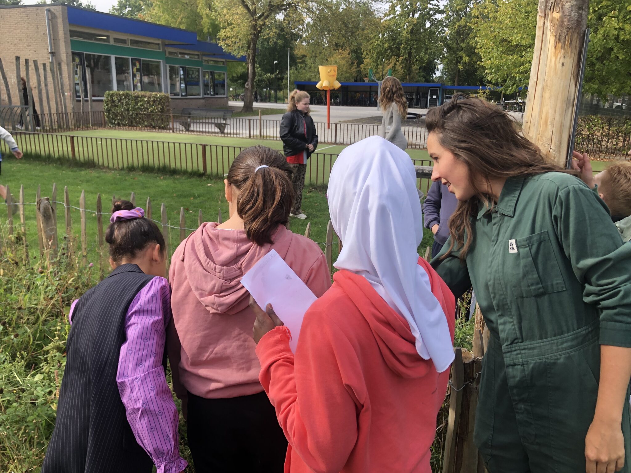 Groep kinderen en een volwassene in overall, observeren iets in een tuin bij een schoolgebouw.