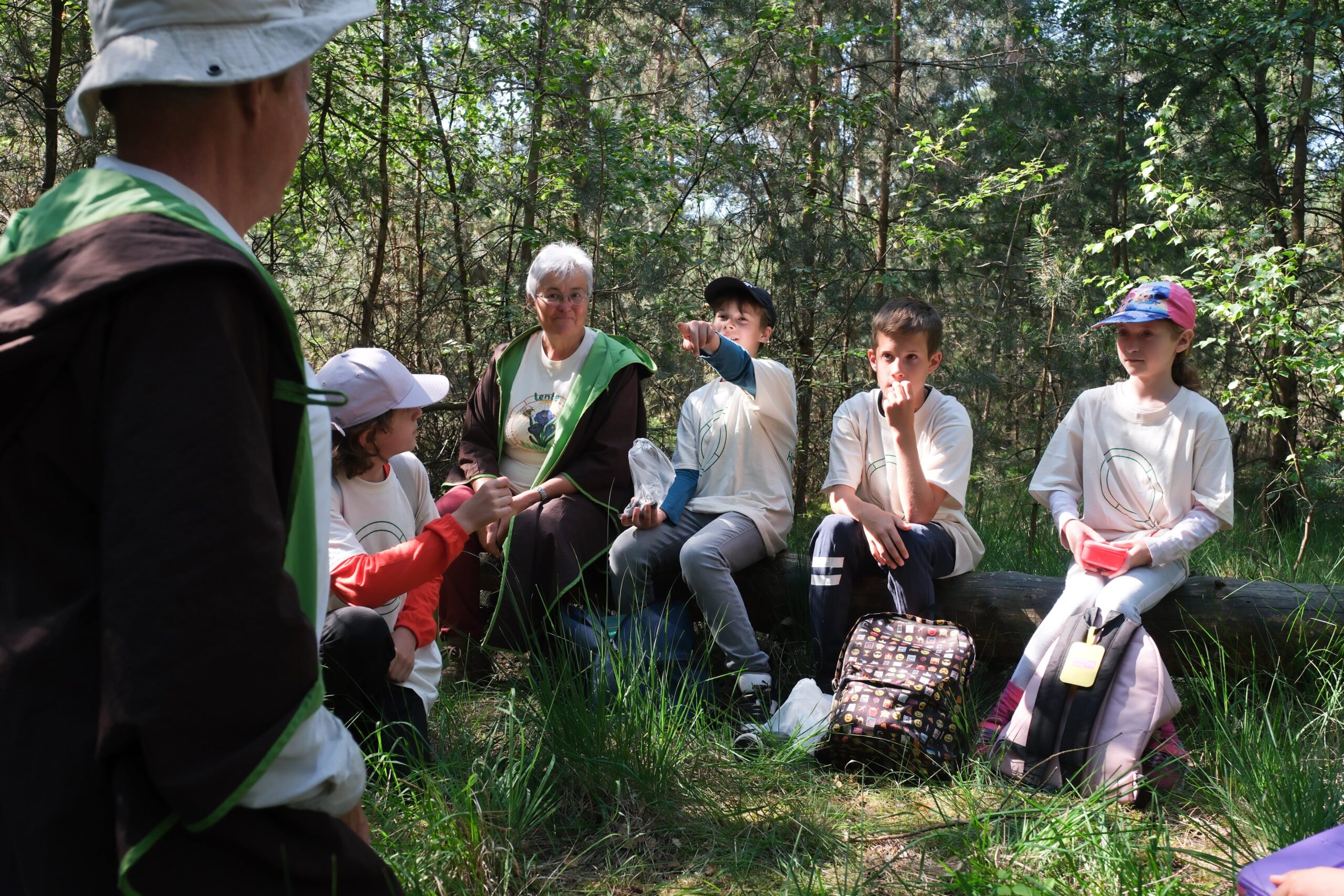 Groep kinderen en een oudere persoon zitten op een boomstam in een bos, praten en ontspannen.