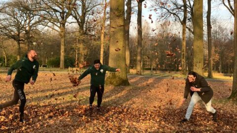 Drie mensen spelen met vallende bladeren in een herfstig bos.