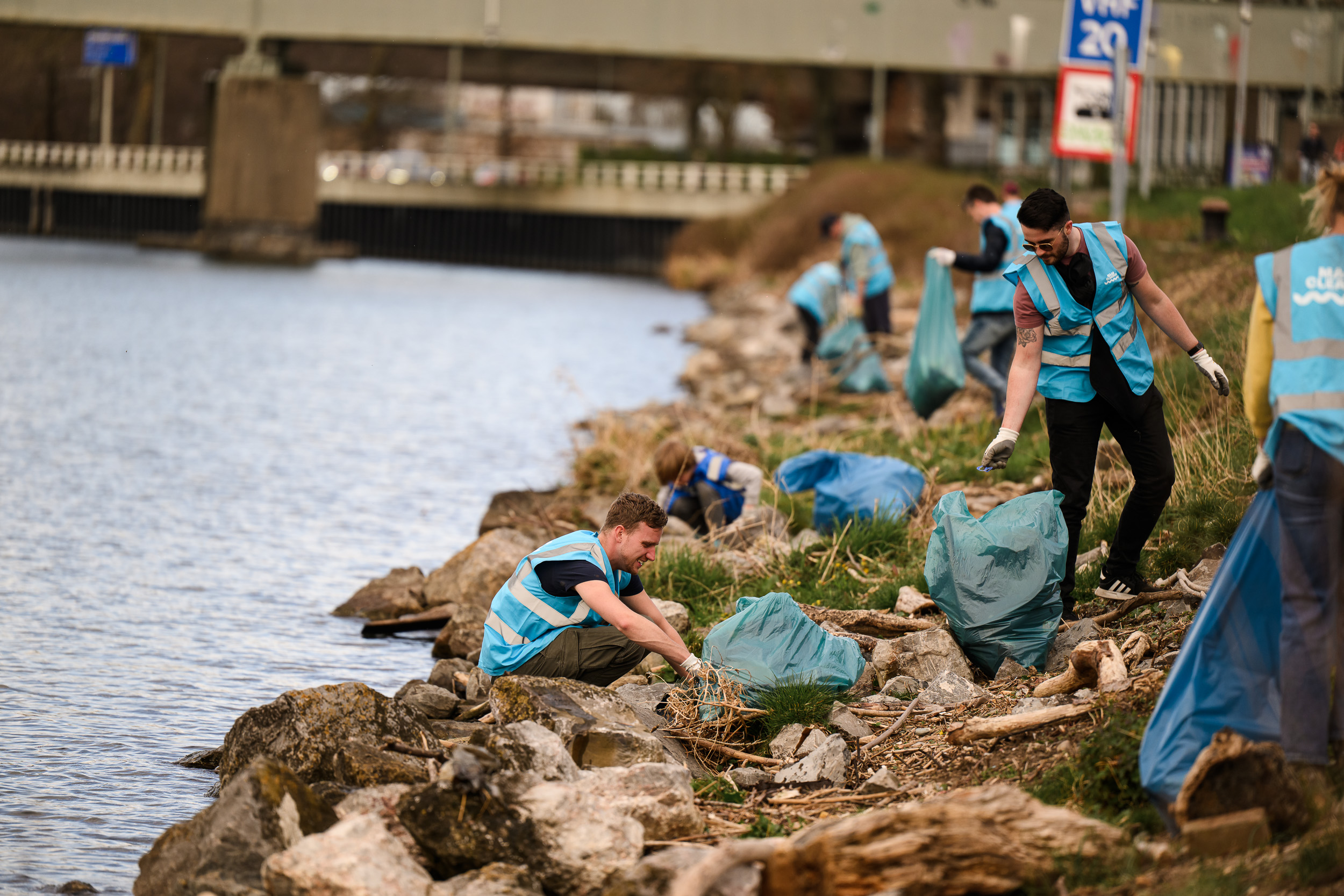 Vrijwilligers ruimen afval op langs een rivier, met blauwe afvalzakken en hesjes.