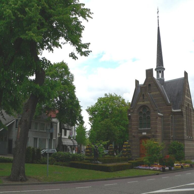 Een kleine kerk met spitse toren, omgeven door bomen en een weg op de voorgrond.