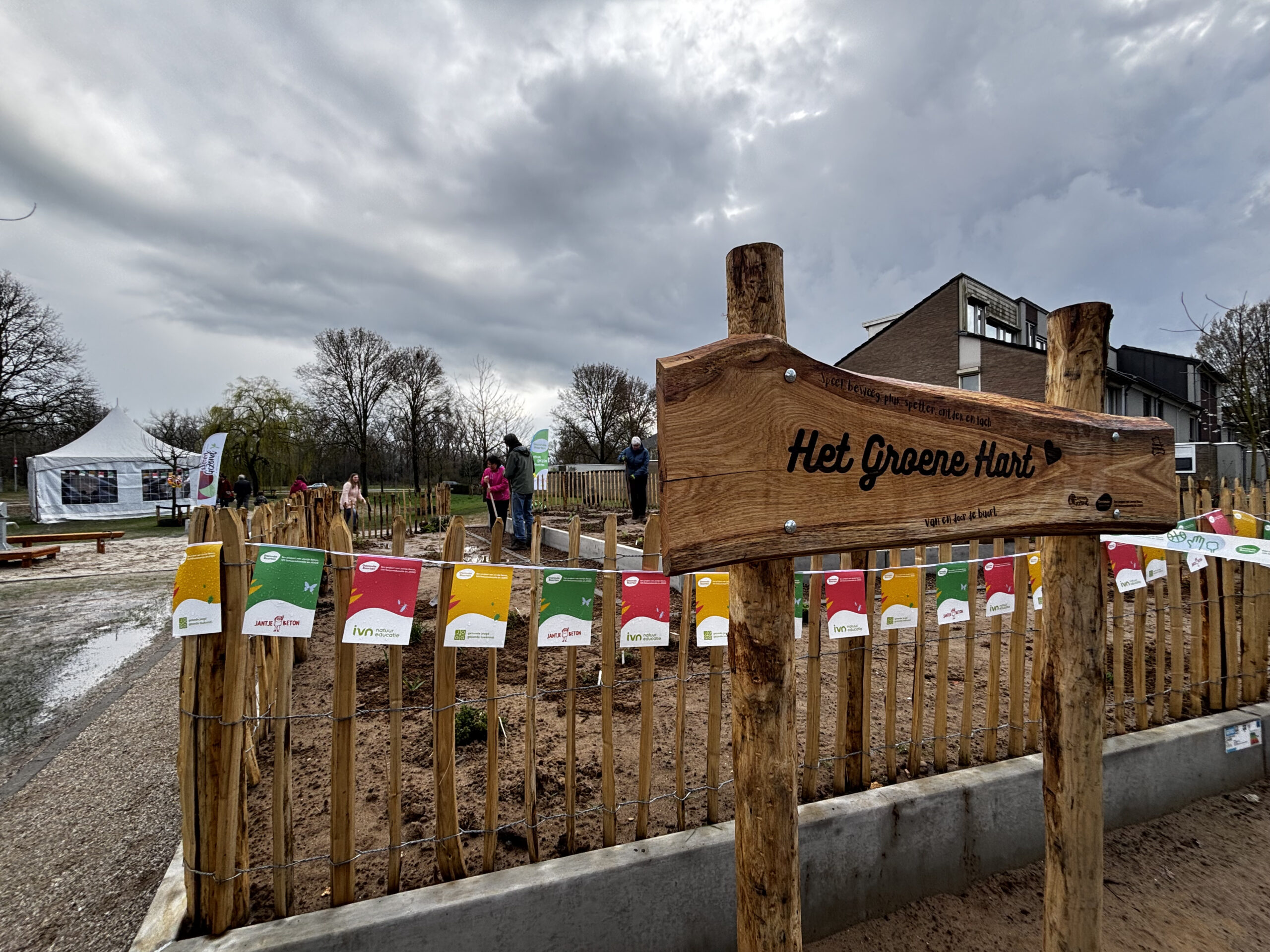Houten bord "Het Groene Hart" bij een omheind tuinperk, met mensen en een tent op de achtergrond.