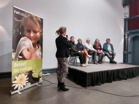 Een vrouw spreekt voor een panel met zes mensen op een podium; naast haar staat een IVN-banner.