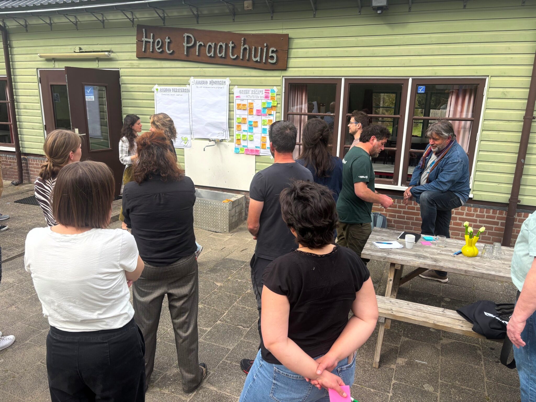 Groep mensen bij "Het Praathuis", kijkend naar een bord met notities buiten.