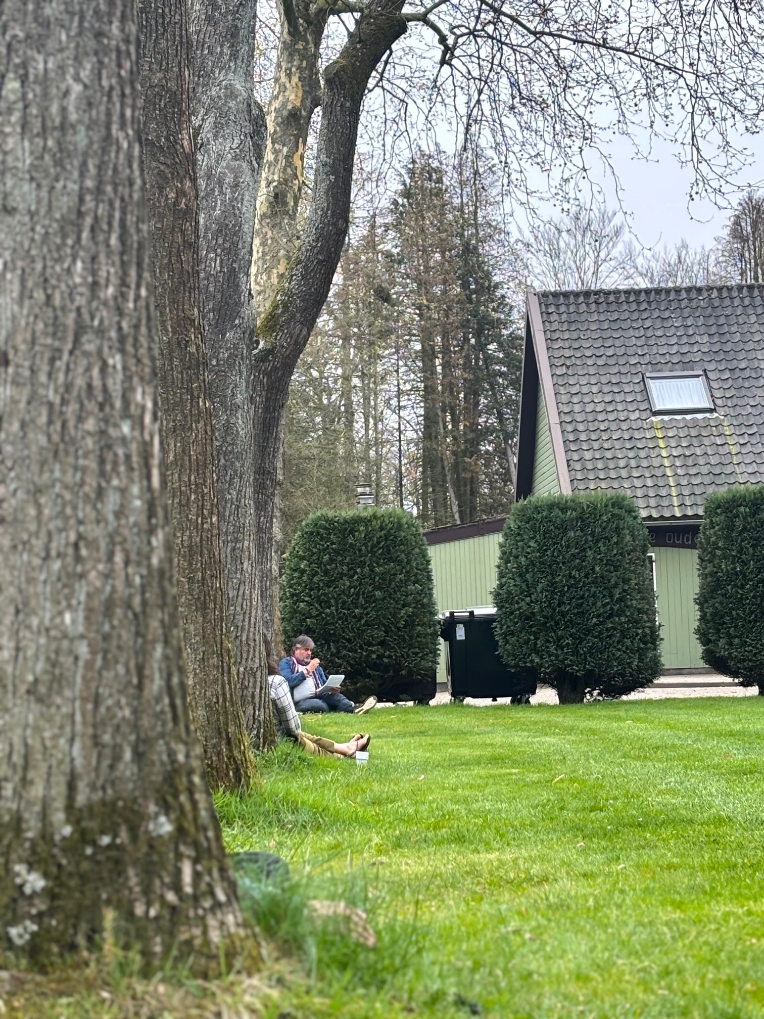 Twee mensen zitten op gras tegen een boom bij een huis met een driehoekig dak.