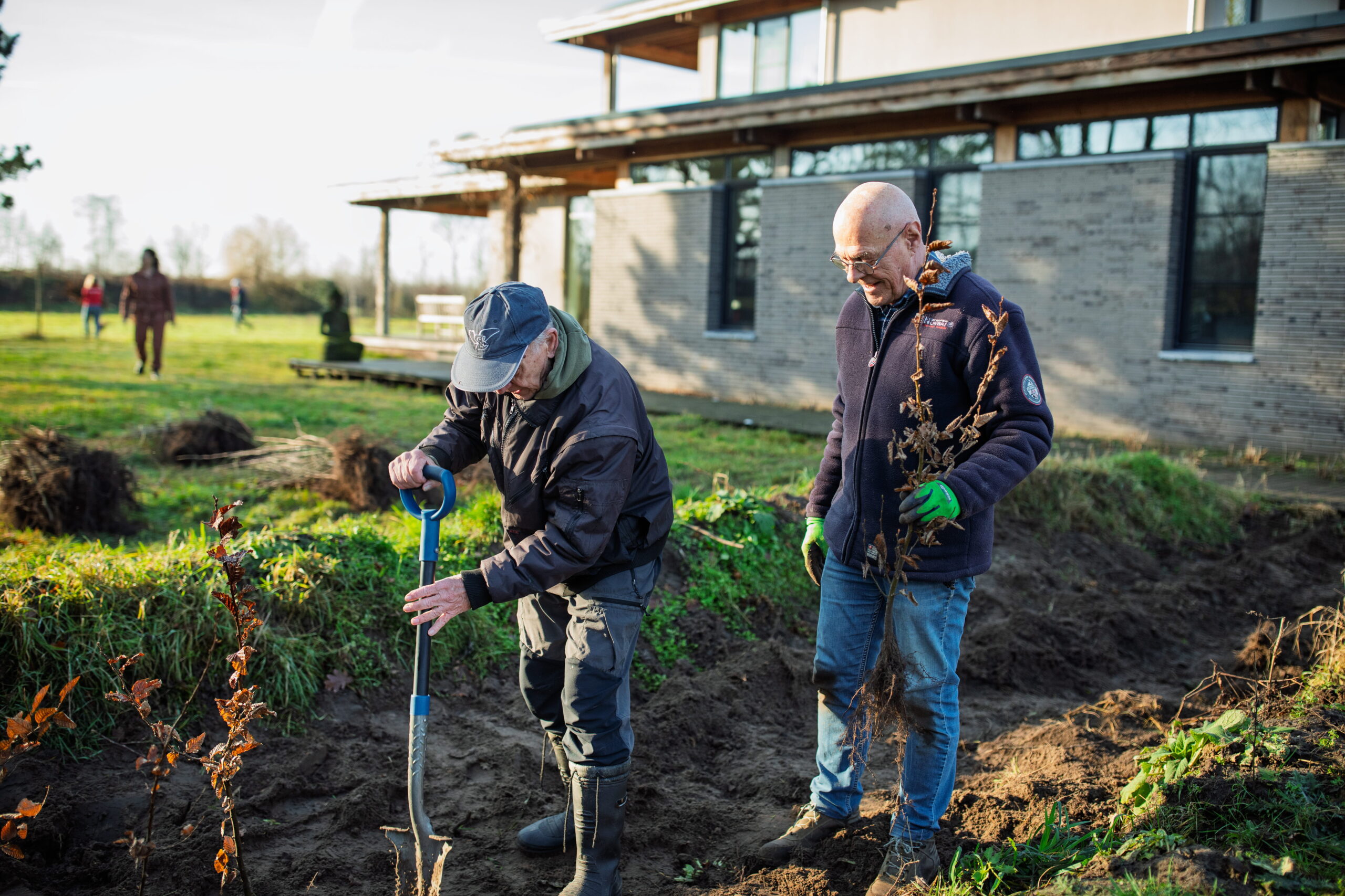Twee mannen planten een boom in de tuin bij een modern gebouw.