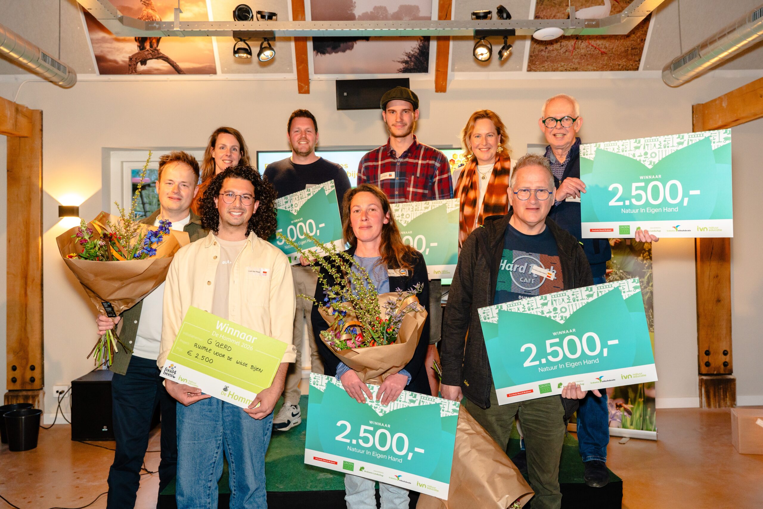 Groep mensen poseert met bloemen en cheques voor een prijsuitreiking, blije gezichten en vreugde.