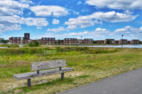 Houten bankje in park met gras, appartementen en blauwe lucht met wolken.