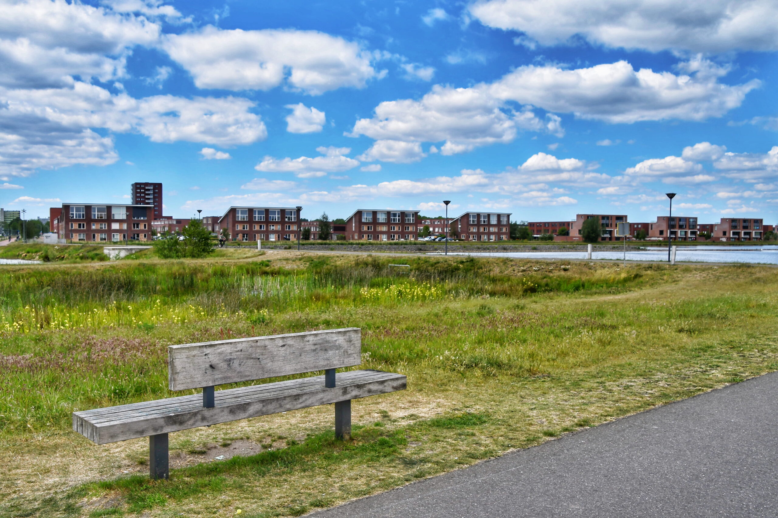 Houten bankje in park met gras, appartementen en blauwe lucht met wolken.