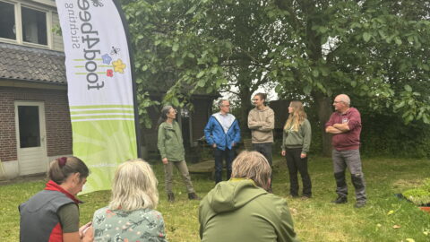 Mensen staan in een tuin naast een Greenpeace-vlag, pratend onder een grote boom.