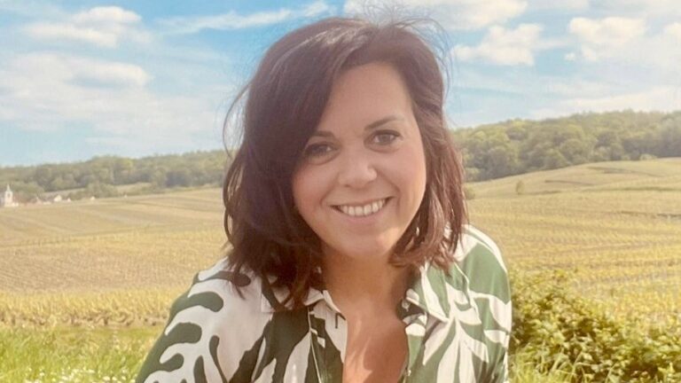Vrouw met donker haar lacht in een groen landschap met velden en blauwe lucht.
