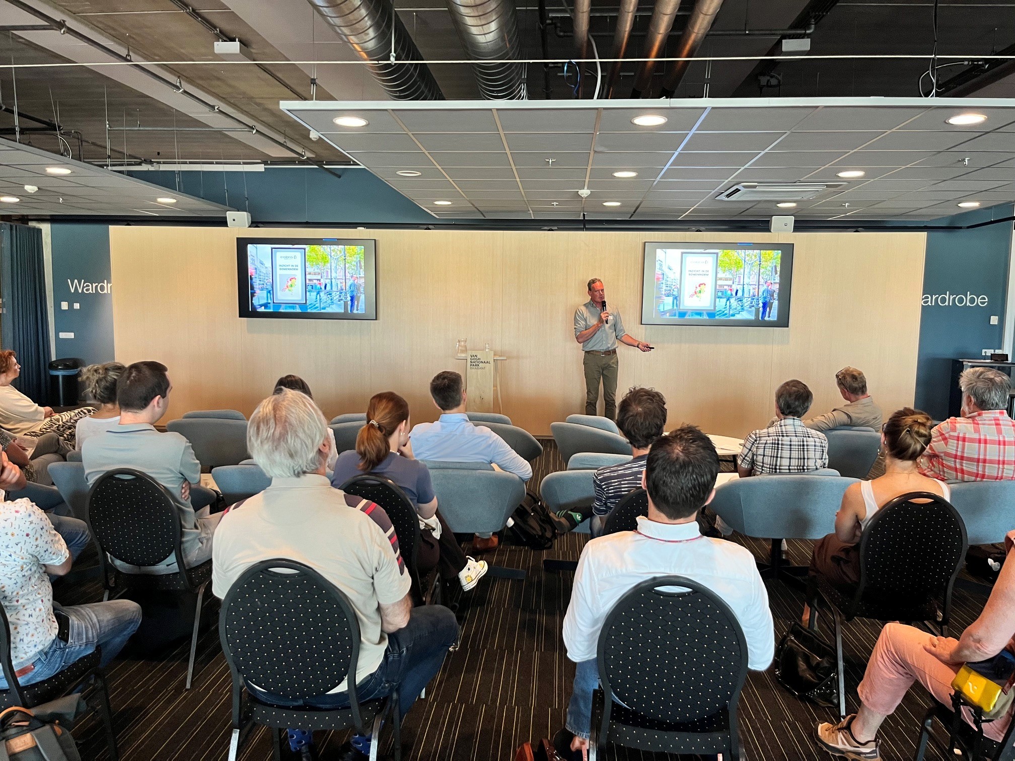 Presentatie met publiek, spreker wijst naar projectie in vergaderruimte.