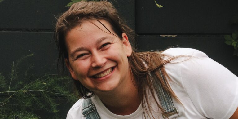 Glimlachende vrouw in tuin, witte shirt en tuinbroek, met groene achtergrond.