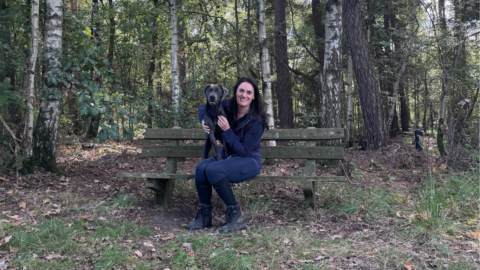 Vrouw zit met hond op een houten bank in een bosrijke omgeving.