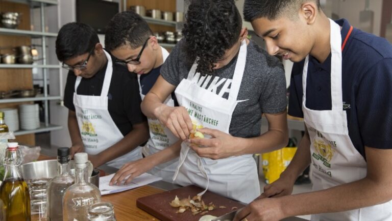 Vier jonge mensen schillen groenten in een keuken met schorten aan.