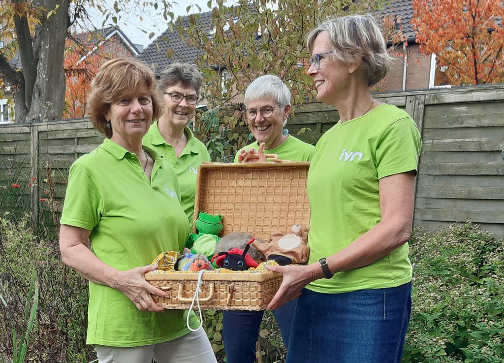 Vier mensen in groene shirts met IVN-logo, bekijken een mand met educatief speelgoed in een tuin.