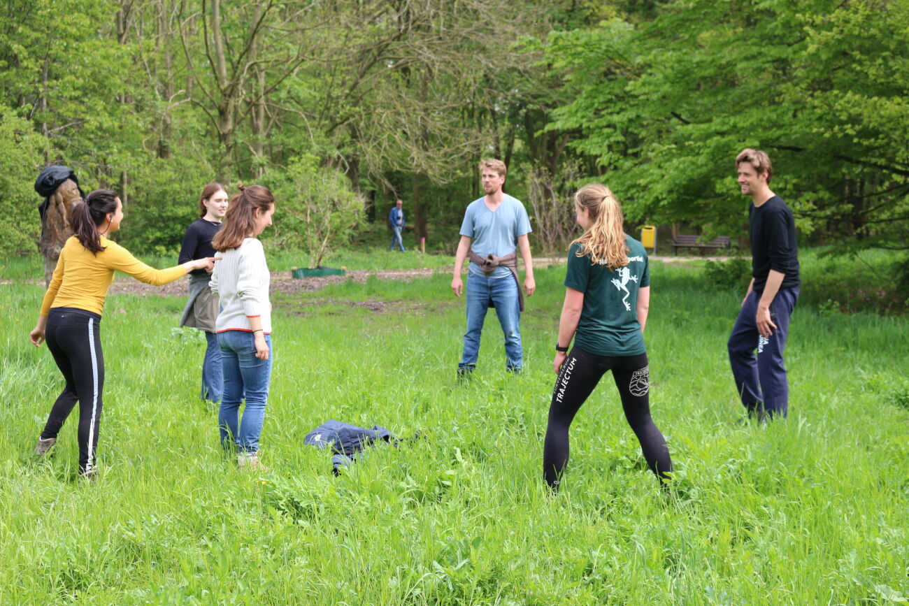 Mensen spelen een buitenspel op gras, omringd door bomen in een bosrijke omgeving.