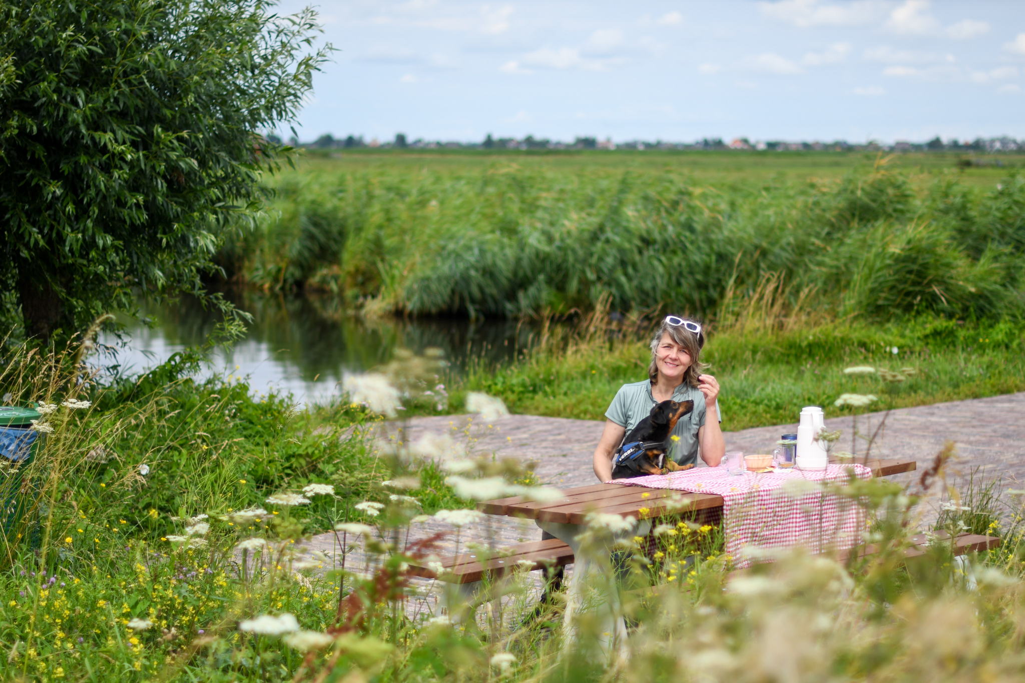 Vrouw zit met hond aan picknicktafel bij kanaal, omringd door groen landschap.