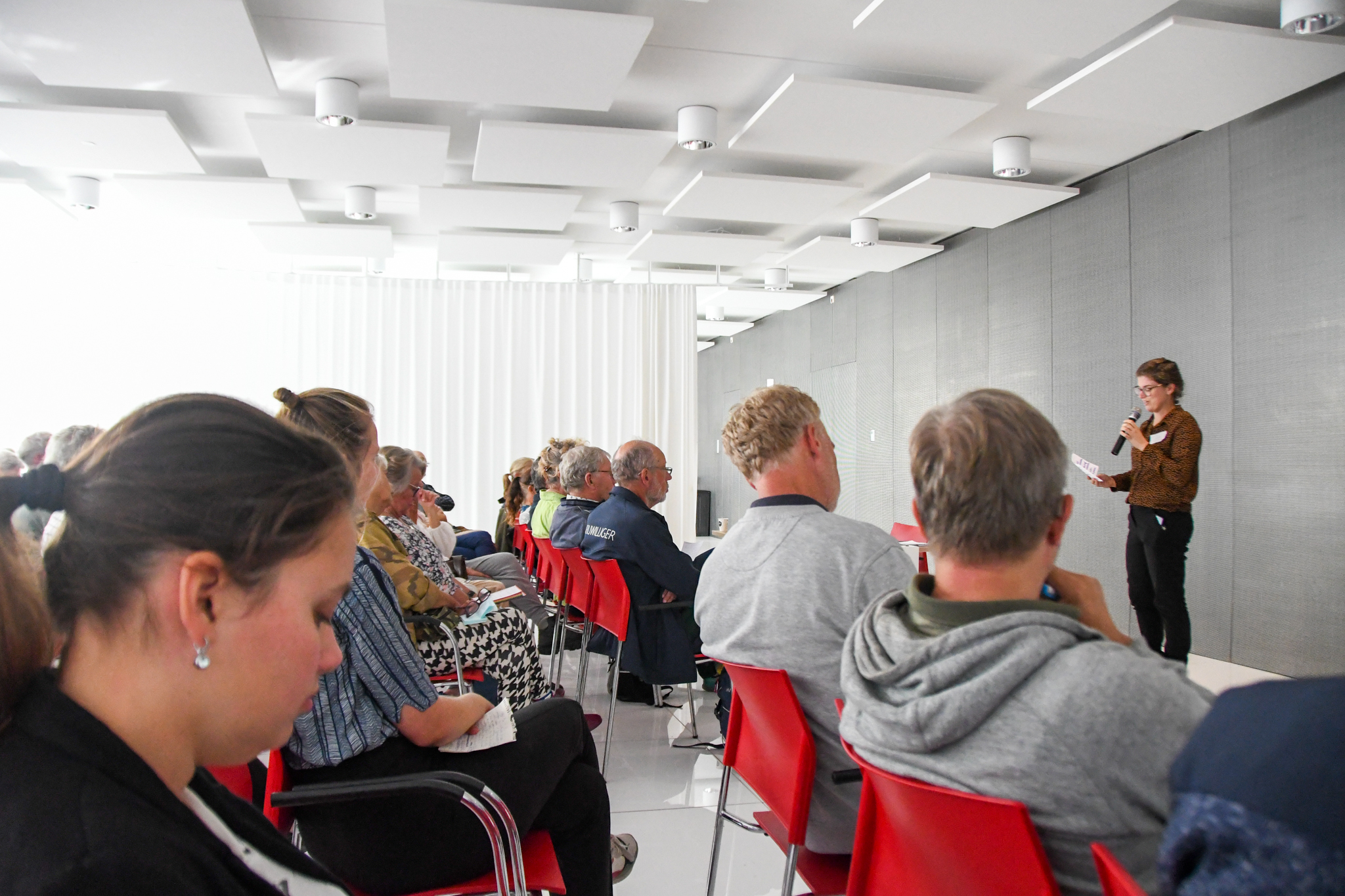 Mensen zitten in een zaal terwijl een spreker met een microfoon spreekt.