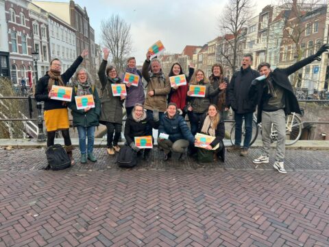 Groep mensen, lachend en poserend met kleurrijke certificaten op een stadsbrug.