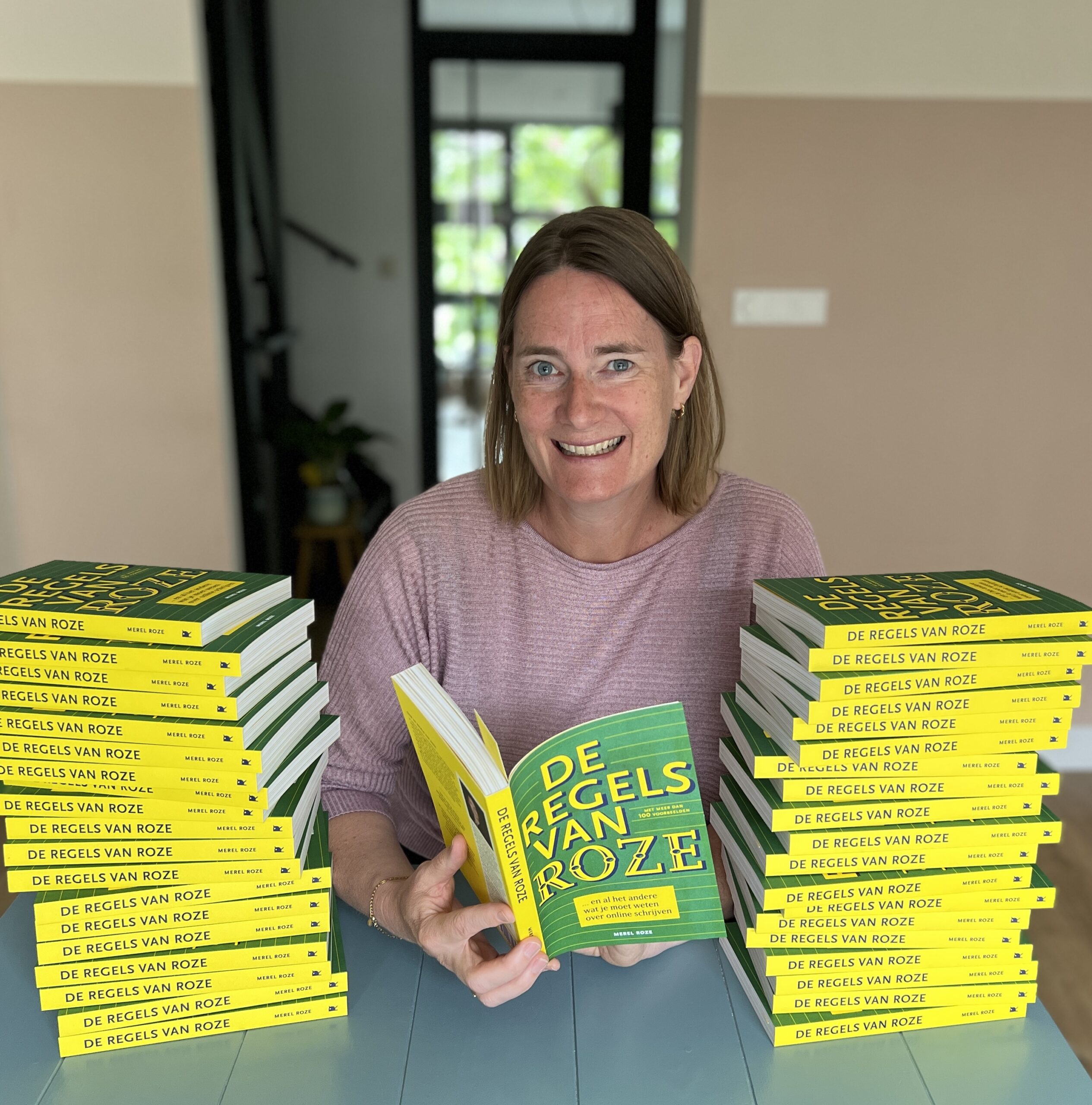Een vrouw poseert lachend met stapels boeken "De Regels van Roze" op een tafel.