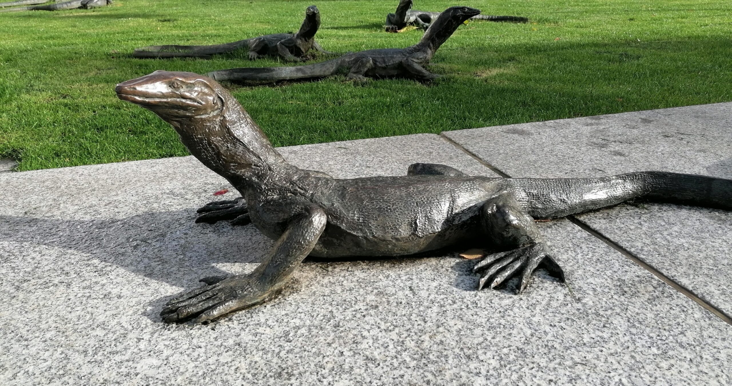 Bronzen leguanenbeelden op stenen trottoir naast grasveld.