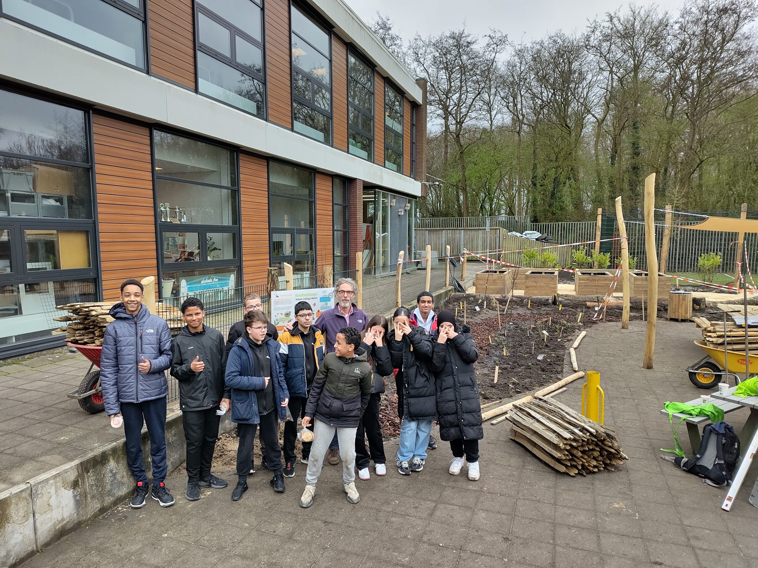 Groep mensen poseert bij een schooltuin in aanbouw naast een schoolgebouw.