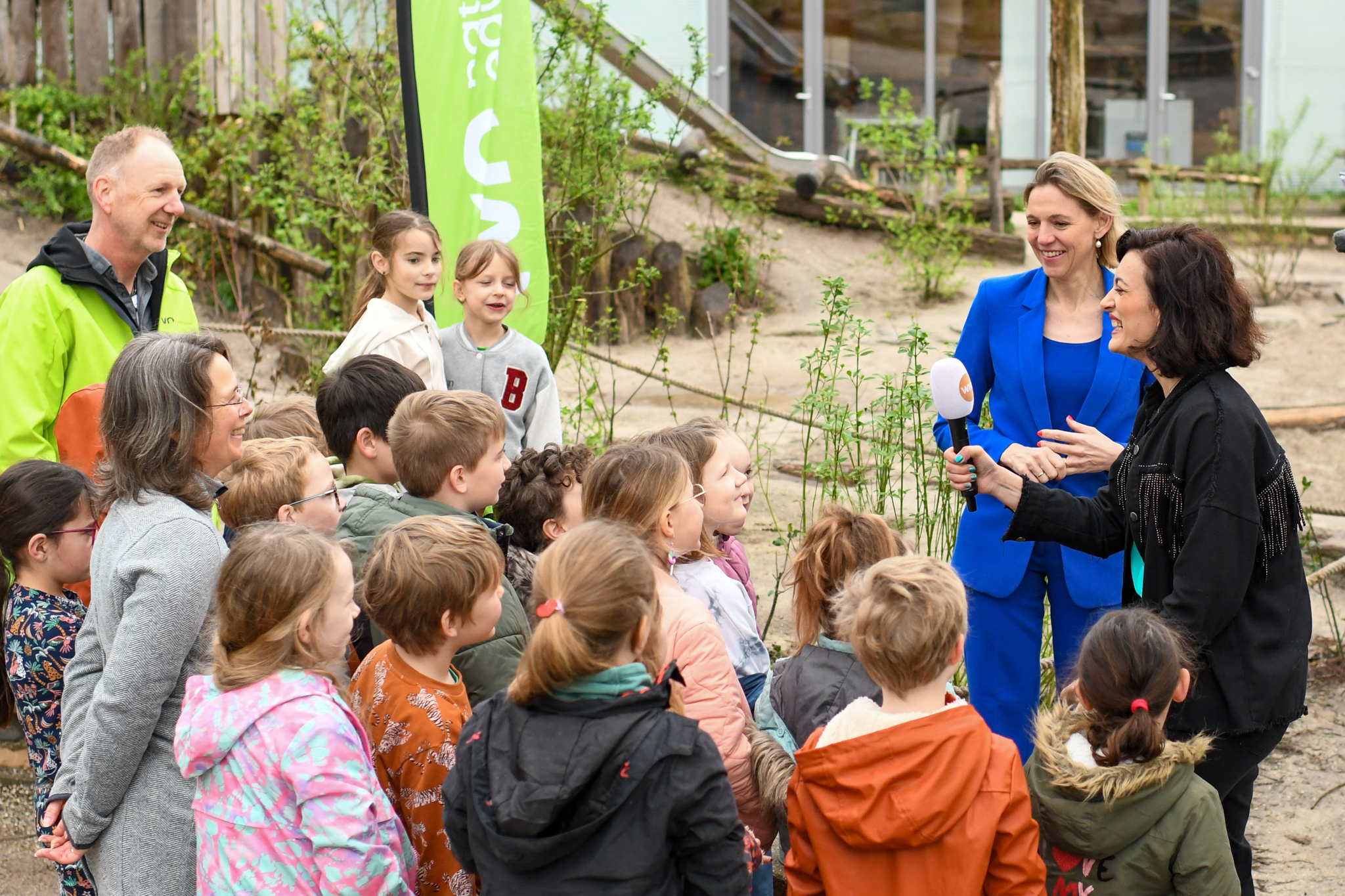Groep kinderen en volwassenen buiten in gesprek met een verslaggever met microfoon.