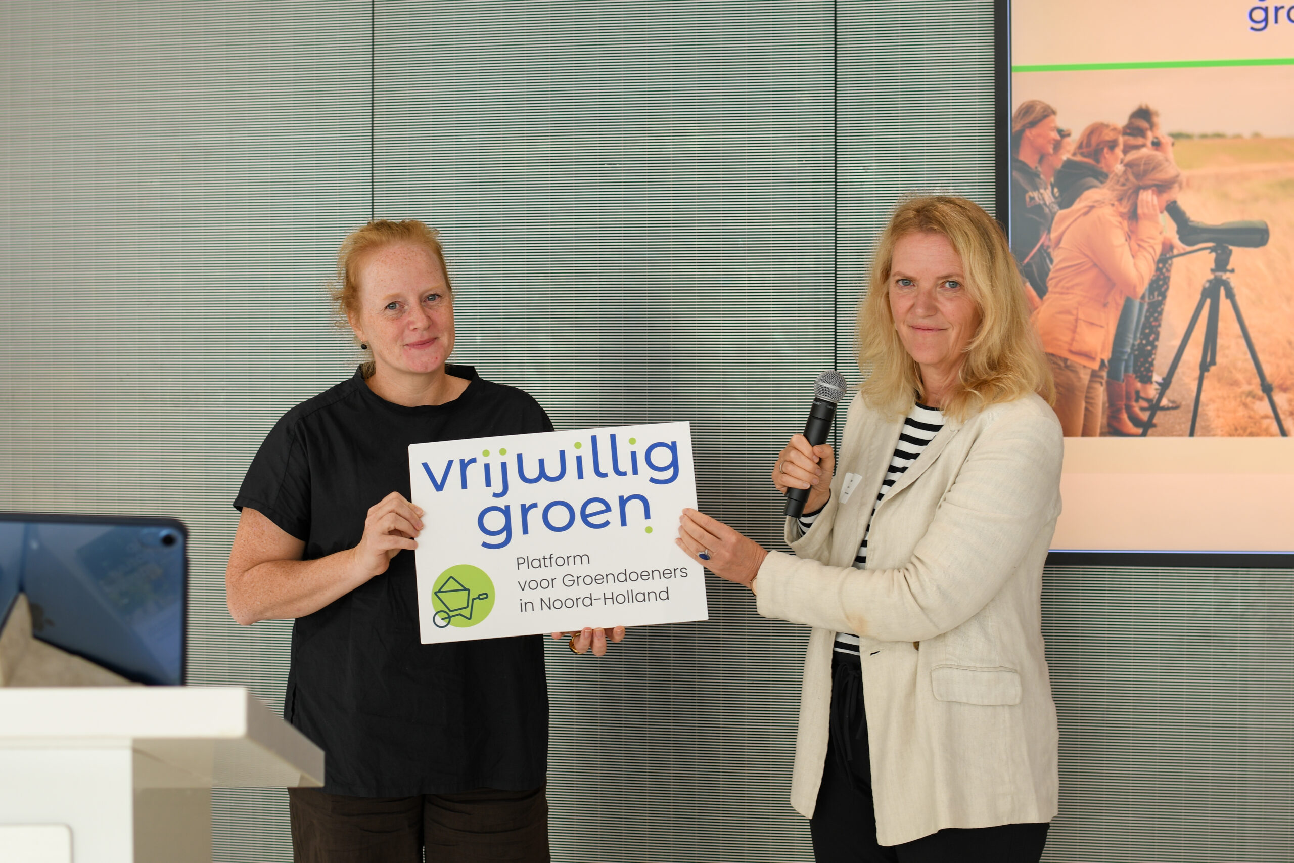 Twee vrouwen houden een bord vast met "vrijwillig groen" erop, tijdens een presentatie.