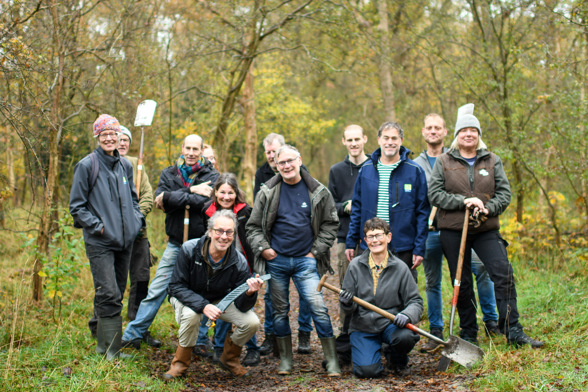 Groep mensen met gereedschap, poseren in een bosrijke omgeving, klaar voor natuurbehoudswerk.