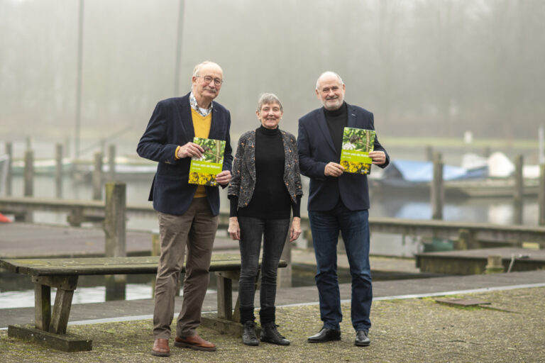 Drie mensen poseren buiten met natuurboeken bij een mistige haven.