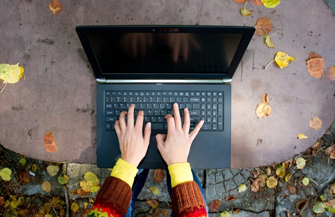 Twee handen typen op een laptop buiten op een tafel met herfstbladeren.